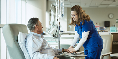 Smiling nurse comforts patient sitting in chair while receiving immunotherapy treatment.