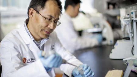 Mingjiang Xu, PhD holds a pipette, concentrating on a sample in a laboratory filled with scientific equipment.