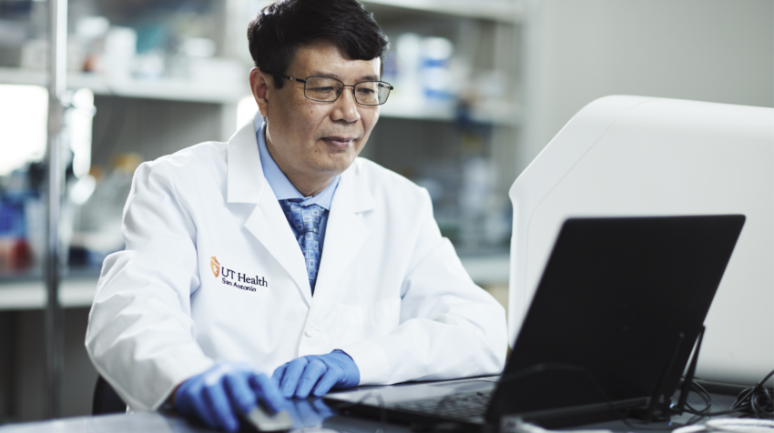 Daohong Zhou, MD researches in a white lab coat works at a laptop in a laboratory, focused and engaged in his task.