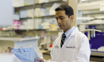 Majeet Rao, PhD focuses, while wearing a white lab coat, examines a document in a laboratory filled with shelves of supplies.