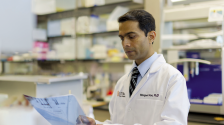 Majeet Rao, PhD focuses, while wearing a white lab coat, examines a document in a laboratory filled with shelves of supplies.