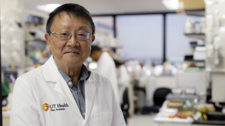 Patrick Sung, DPhil, in a white lab coat stands smiling in a busy laboratory filled with equipment and other researchers in the background.