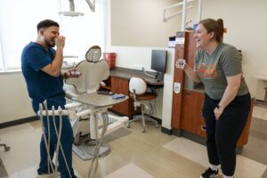A student in blue scrubs laughs while a dental employee holds up a card astonished at the end of a magic trick.