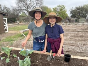 An elderly mother and her daughter wear sun hats and stand next to a raised garden bed.