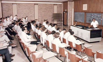 THEN - In a traditional classroom setting in the 1970s, a professor lectures to students who are wearing their white coats.