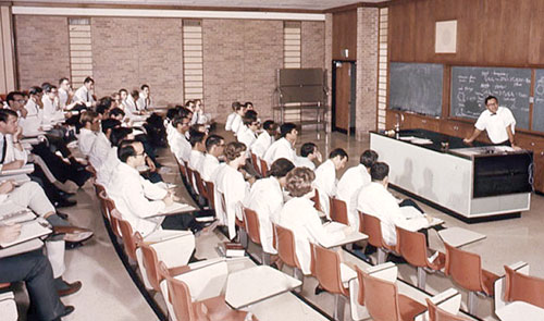 THEN - In a traditional classroom setting in the 1970s, a professor lectures to students who are wearing their white coats.