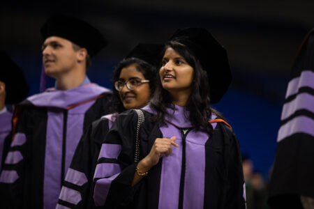 Two female and one male graduate look over their shoulders while walking into the commencement event.