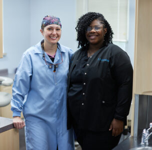 A dentist in a surgeon's cap and a blue dental gown stands next to her assistant in black scrubs. Both are smiling into the camera.