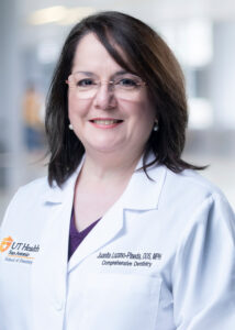 A professional head shot of a female dentist with dark hair and rimless glasses smiles while wearing a white lab coat.