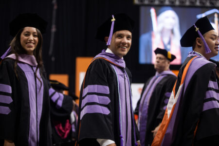 Two male and one female graduates wearing their cap and gowns smile as they enter the commencement event.