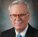 Man with white hair and mustache wearing a black suit, light blue tie and tortoise shell glasses smiles.