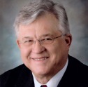 Man with salt and pepper hair wearing a black suit and glasses smiles.