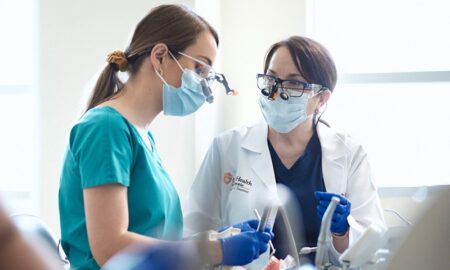 A dental student in green scrubs wears a face mask, dental loupes and gloves while practicing her clinical hand skills on a manikin. Her instructor, in a white lab coat, sits to her right.