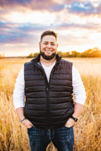 Christian Doll poses in a field smiling with his hands in his pockets.