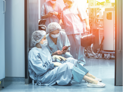 Two nurses in full personal protective equipment sit exhausted in a clinic hallway.